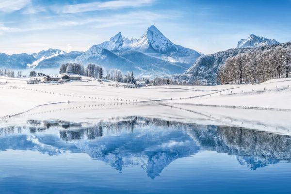 Panoramic view of beautiful white winter wonderland scenery in the Alps with snowy mountain summits reflecting in crystal clear mountain lake on a cold sunny day with blue sky and clouds